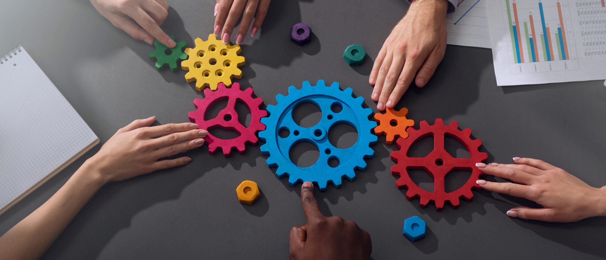 Six hands turning multi-colored spokes and gears on a black table that also has a data sheet and notebook