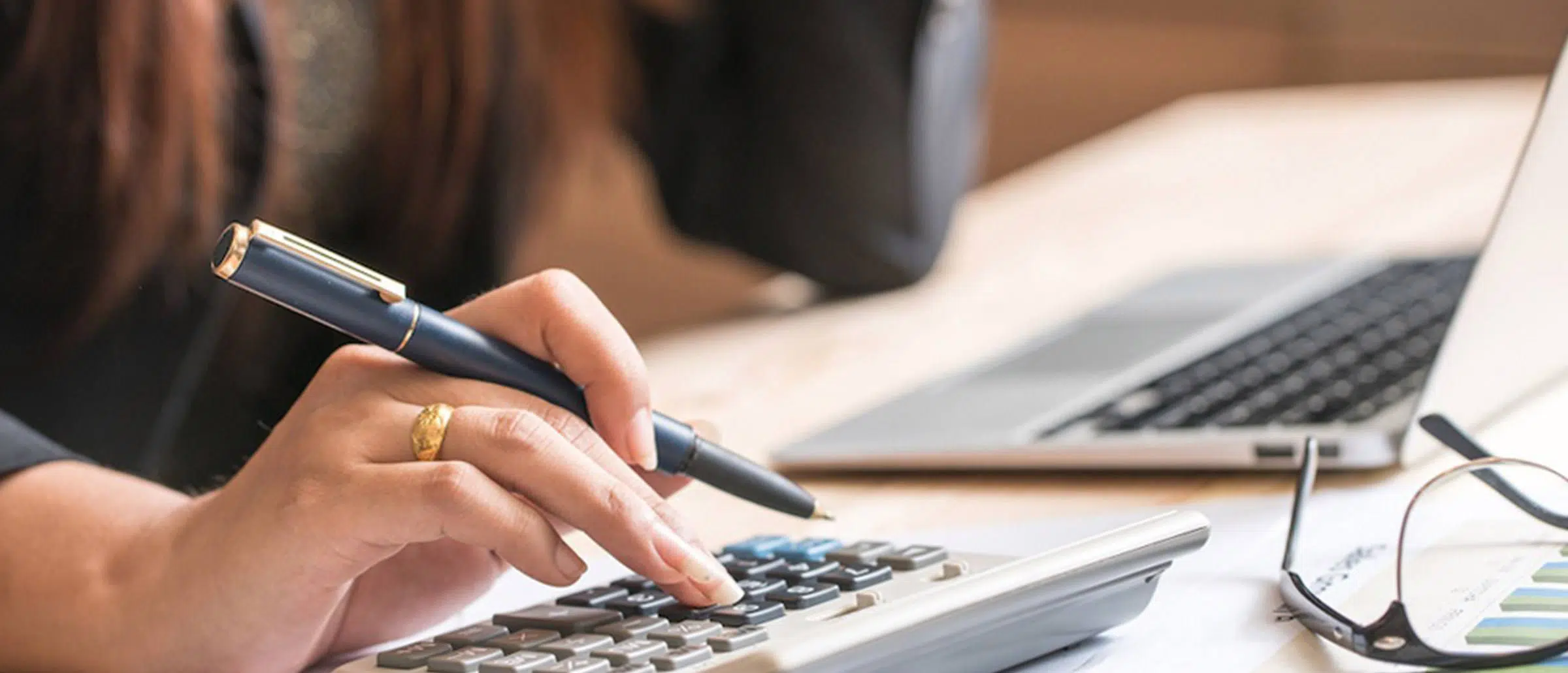 Businessperson pressing keys on a calculator while holding a pen with her glasses and laptop on a table